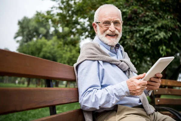 Избирательный фокус улыбающегося пожилого человека с помощью цифрового планшета, сидя на скамейке в парке — стоковое фото