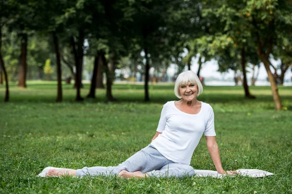 Mujer mayor sonriente relajándose en la esterilla de yoga en el césped verde del parque - foto de stock