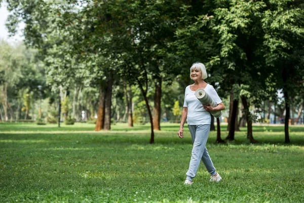 Smiling senior woman holding fitness mat while walking in park — Stock Photo