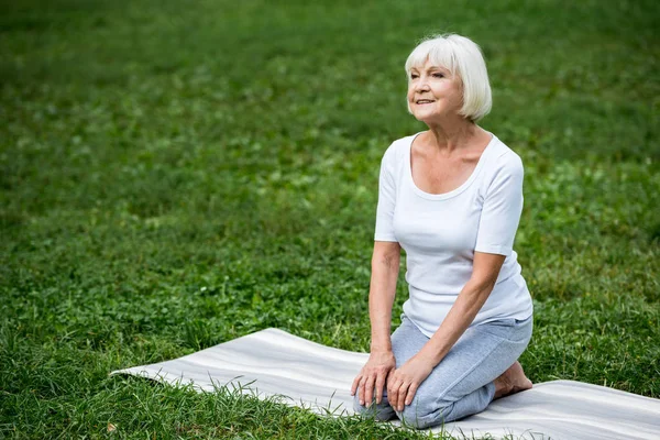 Femme âgée souriante assise sur un tapis de yoga en posture de méditation avec les mains sur les genoux — Photo de stock