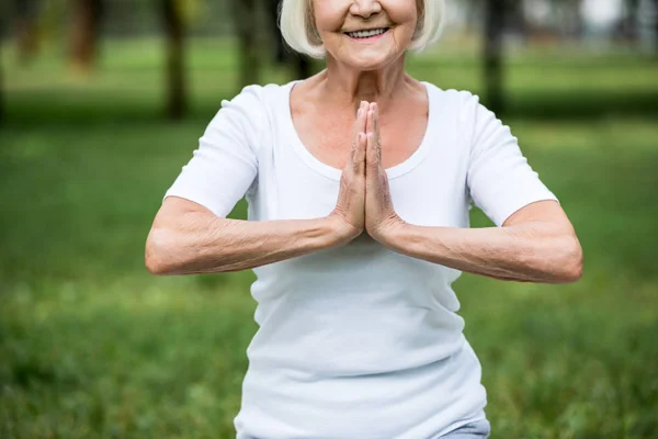 Cropped view of senior woman in meditation sukhasana sitting pose with folded hands — Stock Photo