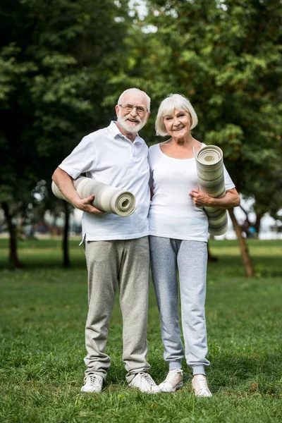 Счастливая пожилая пара обнимается, стоя с фитнес-ковриками в парке — стоковое фото