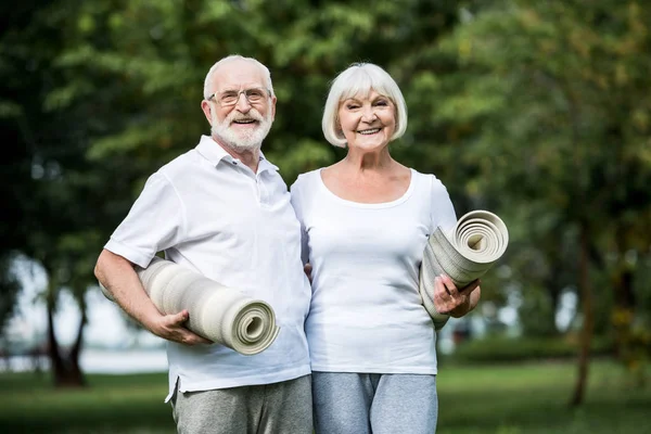Sonriente pareja de ancianos sosteniendo alfombras de fitness mientras está de pie en el parque - foto de stock