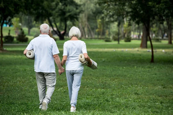 Senior couple holding fitness mats while walking in park and holding hands — Stock Photo