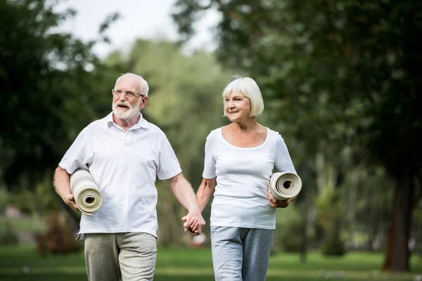 Smiling senior couple with fitness mats walking in park and holding hands — Stock Photo