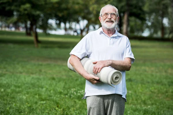 Uomo anziano sorridente che tiene il tappeto di idoneità mentre cammina nel parco — Foto stock