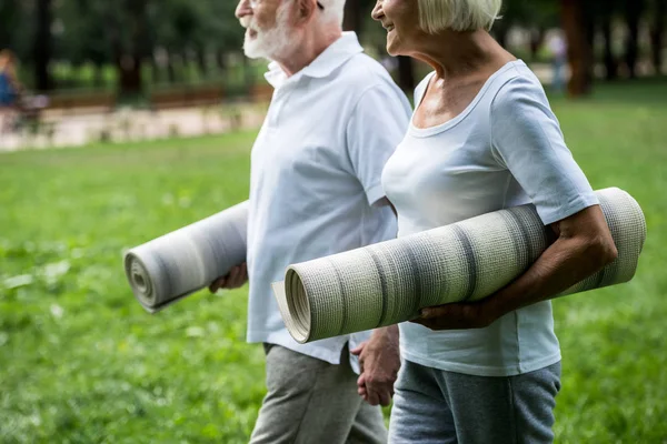 Частичный вид улыбающейся пожилой пары с фитнес-ковриками, гуляющей в парке и держащейся за руки — стоковое фото