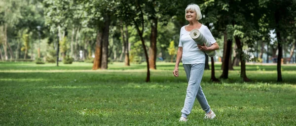 Feliz anciana caminando en el parque y sosteniendo la alfombra de fitness - foto de stock