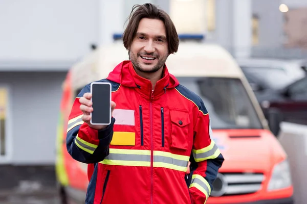 Sonriente paramédico en uniforme rojo sosteniendo smartphone con pantalla en blanco - foto de stock