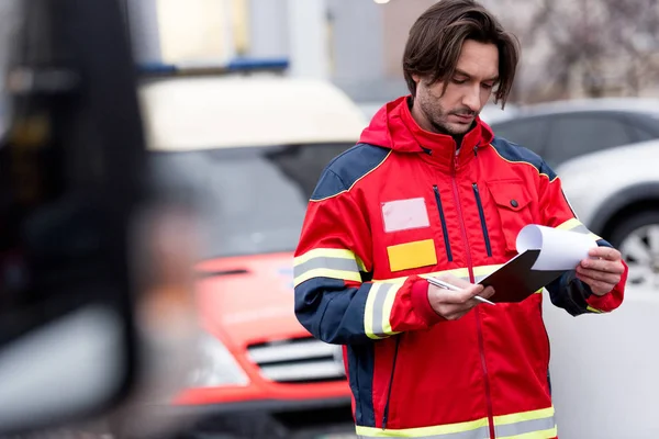 Concentrated paramedic in red uniform writing in clipboard — Stock Photo