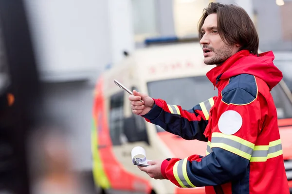 Ambulancier Brunette en uniforme rouge tenant stylo et presse-papiers dans la rue — Photo de stock