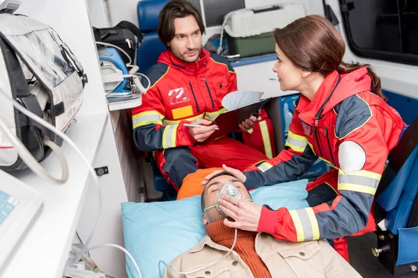 Female paramedic holding oxygen mask on patient while colleague writing diagnosis — Stock Photo