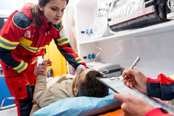 Cropped view of paramedic writing diagnosis while colleague checking pulse — Stock Photo