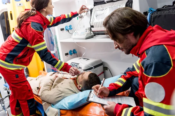 Scrittura paramedica negli appunti mentre il collega tiene la maschera di ossigeno sul paziente — Foto stock