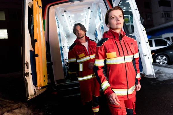 Paramédicos en uniforme rojo de pie cerca de coche ambulancia - foto de stock