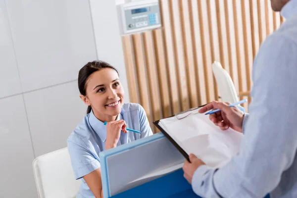 Enfermera sonriente sentada en el lugar de trabajo y hablando con el médico - foto de stock