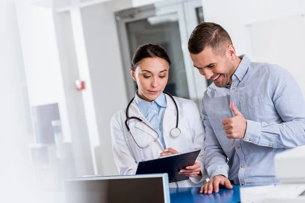 Paciente mostrando el pulgar hacia arriba mientras el médico escribe en portapapeles - foto de stock