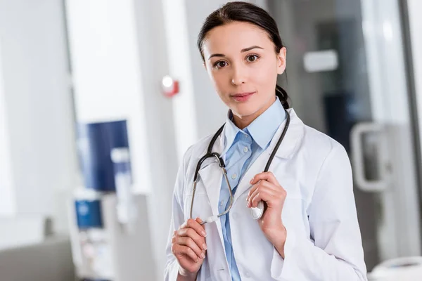 Junge Ärztin im weißen Kittel berührt Stethoskop und blickt in Kamera — Stockfoto