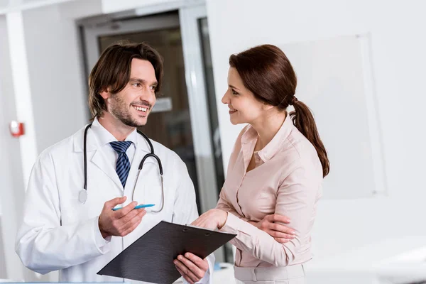 Guapo médico sujetando portapapeles y hablando con el paciente en la clínica - foto de stock