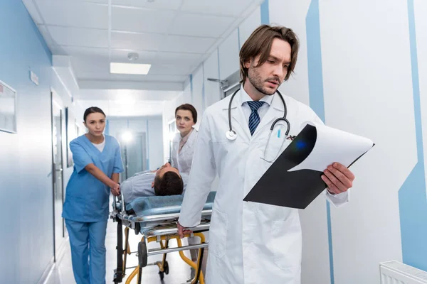 Doctor reading notes while colleagues transporting patient — Stock Photo