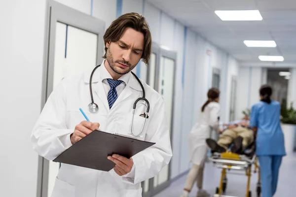Konzentrierter Arzt in weißem Mantel schreibt Notizen in Klemmbrett — Stockfoto