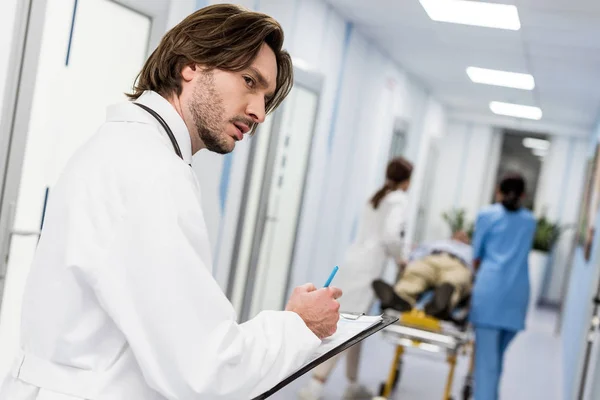 Серьезный молодой врач в белом халате пишет заметки в буфер обмена — стоковое фото