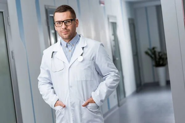 Doctor in glasses and white coat standing with hands in pockets — Stock Photo