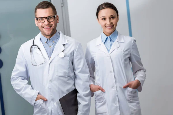 Médicos felices con carpeta y estetoscopio de pie con las manos en los bolsillos - foto de stock