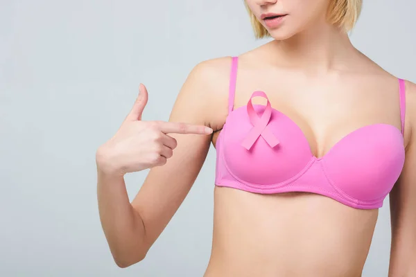 Cropped view of young woman pointing at pink bra with breast cancer awareness ribbon, isolated on grey — Stock Photo
