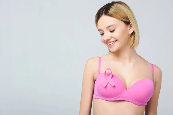 Attraente giovane donna sorridente in reggiseno rosa con nastro consapevolezza cancro al seno, isolato su grigio — Foto stock