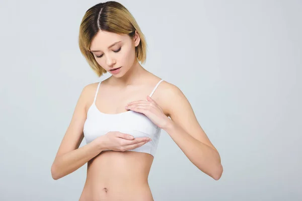 Séduisante jeune femme en sous-vêtements blancs touchant le sein isolé sur gris, concept de cancer du sein — Photo de stock