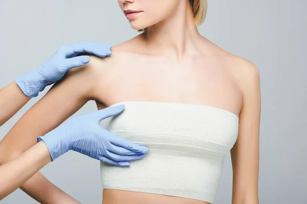 Cropped view of plastic surgeon in latex gloves checking female breast in bandage, isolated on grey — Stock Photo