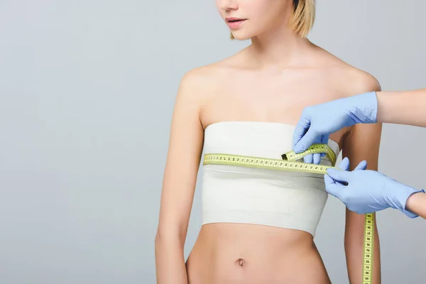 Обрізаний вид лікаря, який тримає вимірювальну стрічку на жіночих грудях з бинтом після пластичної хірургії, ізольовано на сірому — Stock Photo