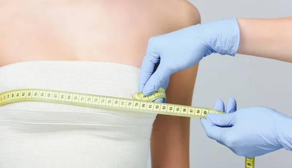 Обрезанный вид пластического хирурга, держащего измерительную ленту на женской груди с повязкой, изолированный на серой — стоковое фото