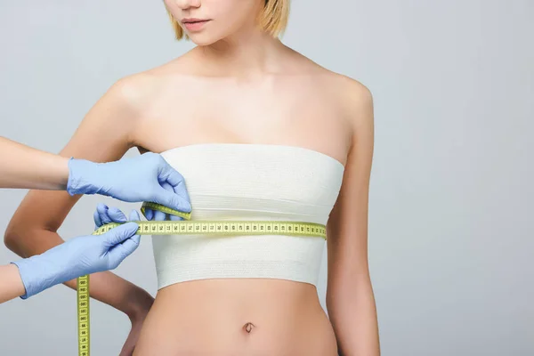 Обрезанный вид пластического хирурга в латексных перчатках, держащего измерительную ленту на женской груди с повязкой, изолированный на сером — стоковое фото