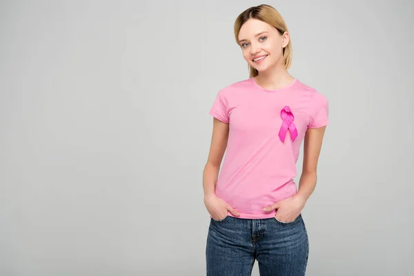 Fille blonde souriante en t-shirt rose avec ruban de sensibilisation au cancer du sein, isolé sur gris — Photo de stock