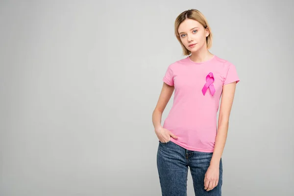Приваблива молода жінка в рожевій футболці зі стрічкою обізнаності про рак молочної залози, ізольована на сірому — стокове фото