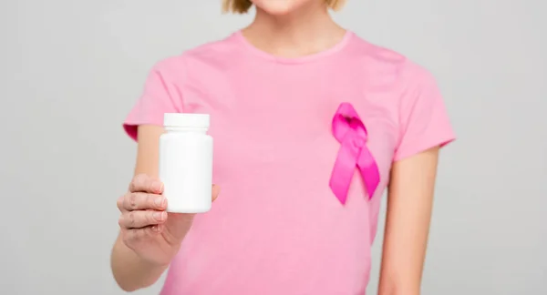 Vue recadrée de la jeune femme en t-shirt rose avec un ruban de sensibilisation au cancer du sein tenant bouteille avec des pilules, isolé sur gris — Photo de stock