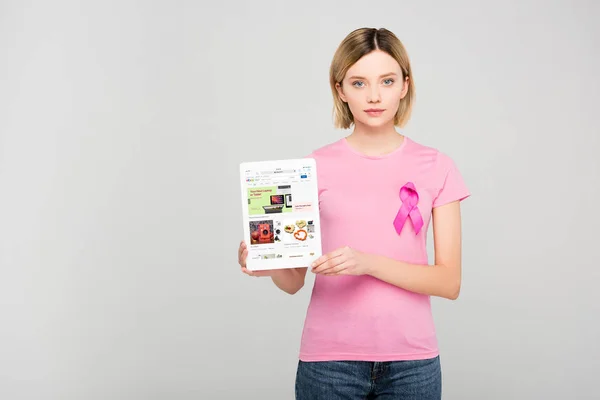 Chica rubia en camiseta rosa con el cáncer de mama cinta de conciencia celebración de la tableta con la aplicación de ebay, aislado en gris - foto de stock