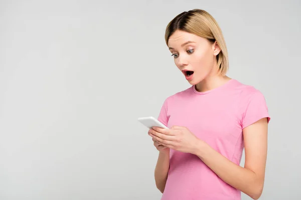 Choqué fille blonde en t-shirt rose en utilisant un smartphone, isolé sur gris — Photo de stock