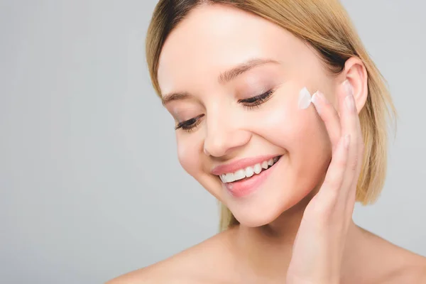 Schöne lächelnde junge Frau, die kosmetische Creme auf Gesicht isoliert auf grau aufträgt — Stockfoto