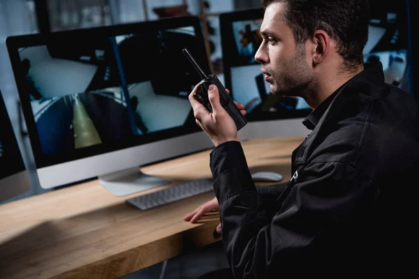 Guapo guardia en uniforme sosteniendo walkie-talkie y mirando el monitor de la computadora - foto de stock