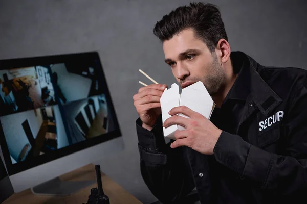 Охранник в форме ест нездоровую пищу и смотрит на монитор компьютера — стоковое фото