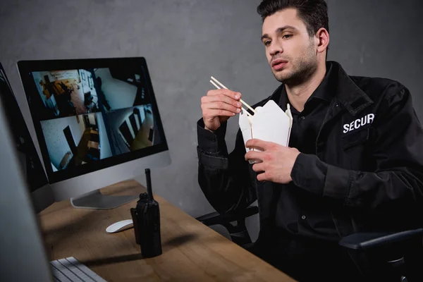 Schöner Wachmann in Uniform, der Essen zum Mitnehmen isst und auf den Computermonitor schaut — Stockfoto