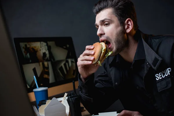 Wachmann in Uniform isst Burger und blickt auf Computermonitor — Stockfoto