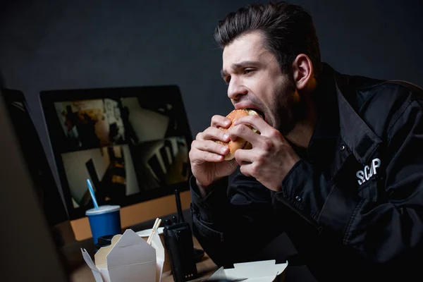 Guarda em uniforme comer hambúrguer e olhando para monitor de computador — Fotografia de Stock