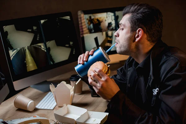 Guard drinking, holding burger and looking at computer monitor at workplace — Stock Photo