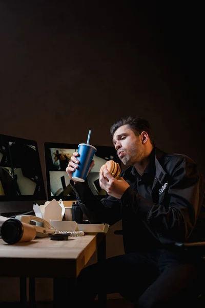 Guapo guardia comiendo hamburguesa y sosteniendo papel taza en el lugar de trabajo - foto de stock