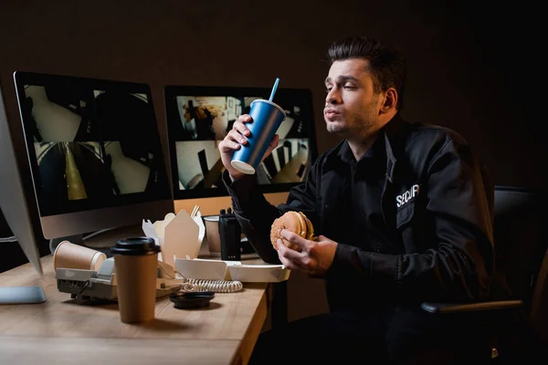 Guarda comer hambúrguer, segurando copo de papel e olhando para monitor de computador — Fotografia de Stock