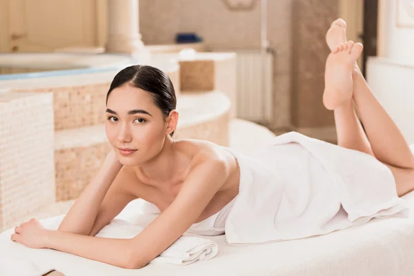 Attractive asian woman in towel lying on massage table and looking at camera — Stock Photo